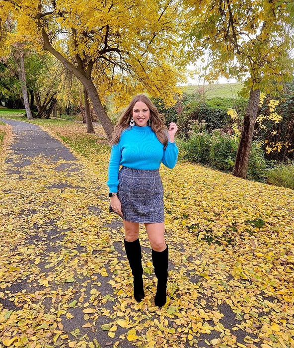 A reviewer poses on a leaf-strewn park trail, wearing a blue sweater, a patterned skirt, and black knee-high boots