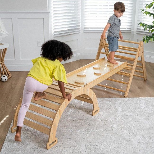 Two children enjoying playtime on a wooden indoor climbing and balancing structure in a bright room with light-colored furniture and a cozy rug
