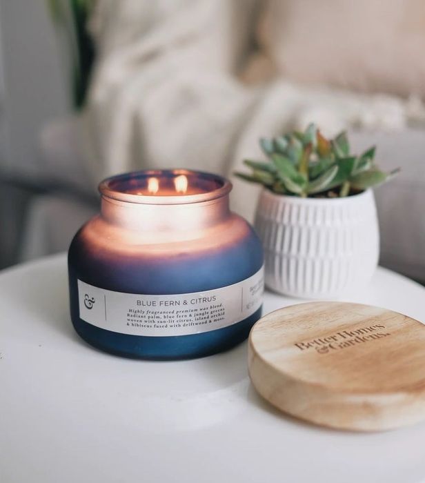 A blue fern and citrus scented candle glowing softly in a blue votive, placed on a table.