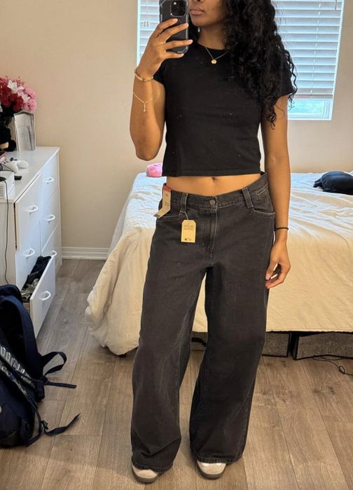 A person poses for a mirror selfie in their black top and wide-leg jeans, standing in a bedroom.