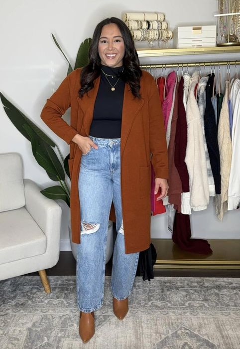 A woman stands smiling in front of a clothing rack filled with various jackets. She is wearing a long brown coat, a black top, and ripped high-waisted jeans with brown shoes.