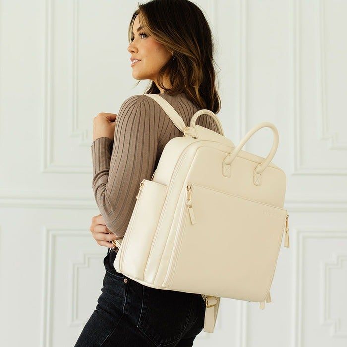 A woman dressed in a ribbed sweater and jeans, carrying a trendy beige backpack, posing against a white wall. Ideal for a fashion-focused shopping guide.