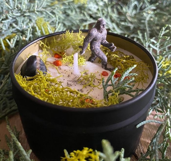A candle featuring a mini Sasquatch figurine with nature-inspired accents on top of the wax.