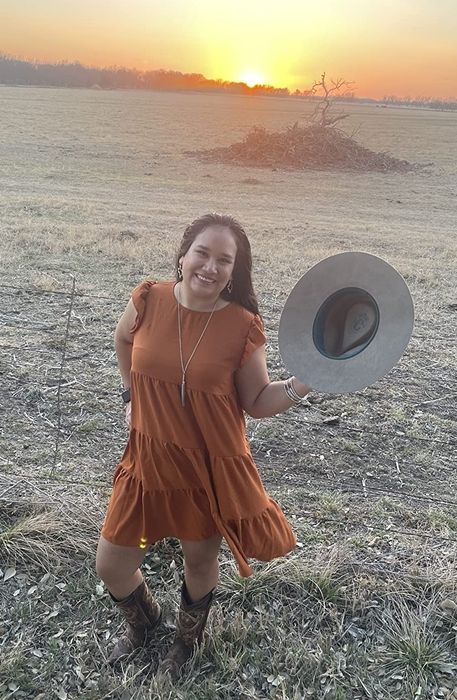 A customer outdoors at sunset, wearing a dress, brown cowboy boots, holding a hat, and smiling