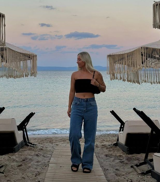 A woman casually dressed in a black top and blue jeans, standing on a beach boardwalk as dusk sets in.