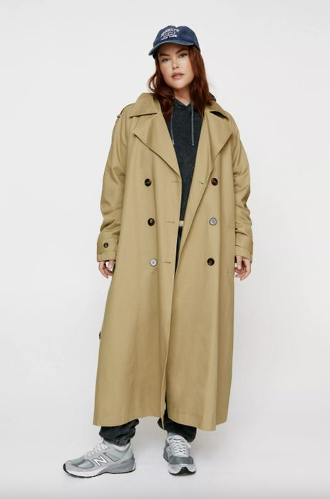 A woman wears a long trench coat over a hoodie, pants, and sneakers, accessorized with a baseball cap, posing in front of a neutral background.