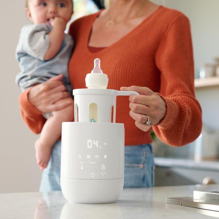 A parent cradles a baby while using a sleek bottle warmer in a kitchen, highlighting its convenience for everyday use.