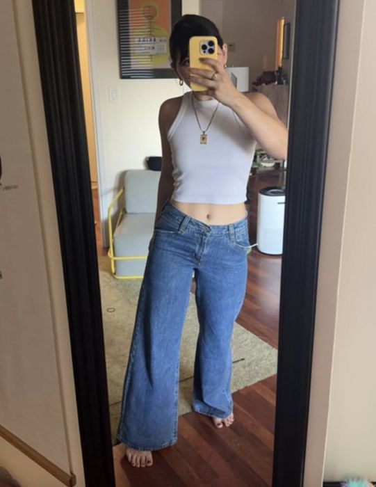 A person wearing a white tank top and blue jeans takes a mirror selfie, with their phone held up to cover their face.
