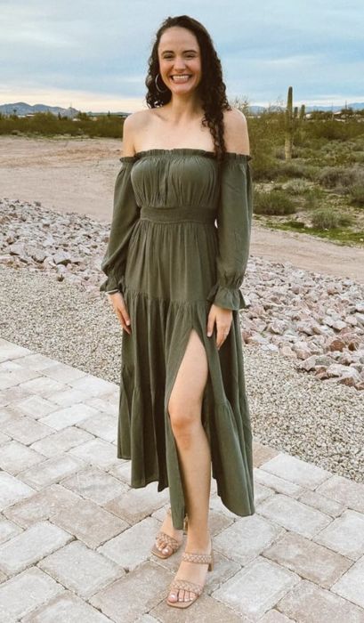 A reviewer strikes a pose outdoors in an off-shoulder, long-sleeved dress featuring a thigh-high slit, standing on a paved path with a desert backdrop.