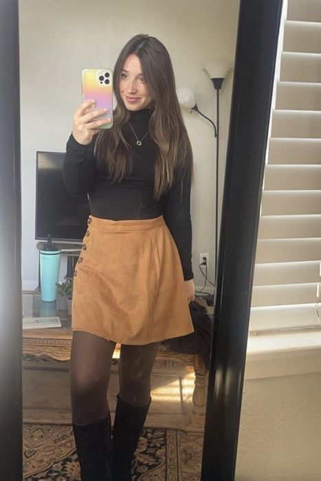 A woman captures a mirror selfie in a room, dressed in a black top, tan skirt, black tights, and boots. Her phone case is bright and colorful.