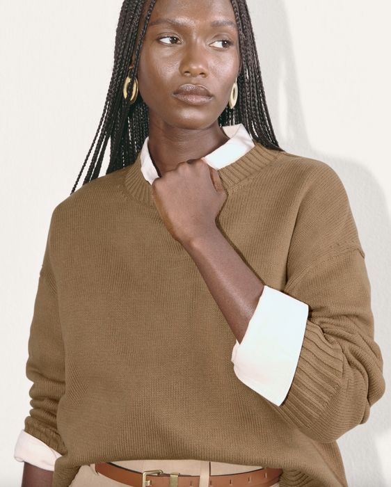 A person with long braided hair and hoop earrings wears a white collared shirt beneath a ribbed sweater, looking thoughtfully to the side.