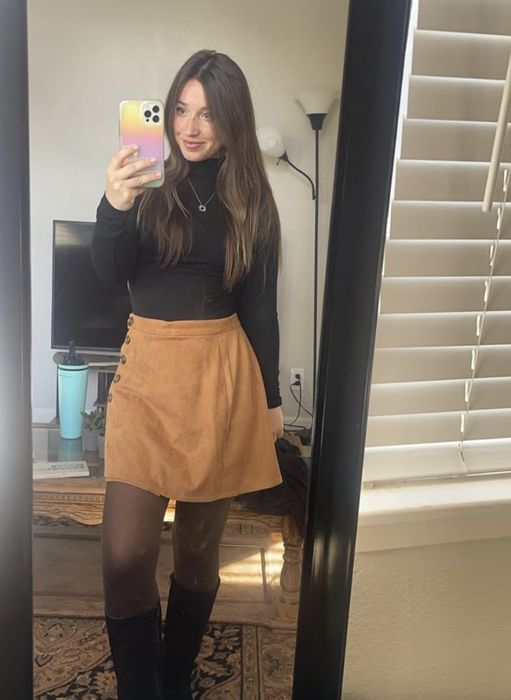 A woman snaps a mirror selfie in a room. She sports a black top, a tan skirt, black tights, and boots. Her phone case is colorful.