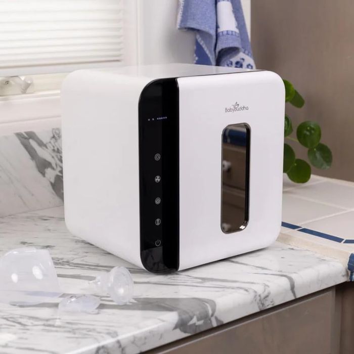 A BabyBuddha sterilizer sits on a marble countertop, featuring control buttons with a breast pump part placed nearby.