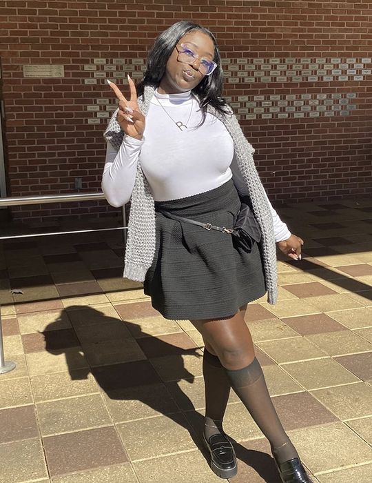 A person strikes a playful pose, flashing a peace sign. They are dressed in a white turtleneck, a gray knitted vest, and a black skirt paired with knee-high socks and loafers.