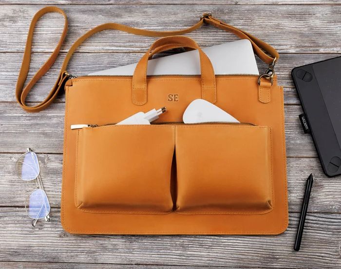 A leather laptop bag with dual front pockets, packed with a charger and mouse. Inside, you’ll find glasses, a pen, a tablet, and a neatly folded laptop—all ready to go.