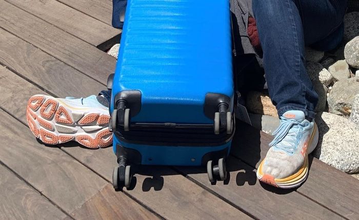 A reviewer sitting on the ground next to a blue suitcase, wearing sports shoes.
