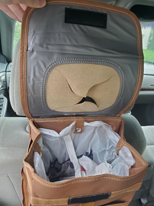 A portable car trash bin designed with adjustable straps, a mesh top opening, and internal lining to keep waste contained. It stays open thanks to a supportive hand inside the vehicle.