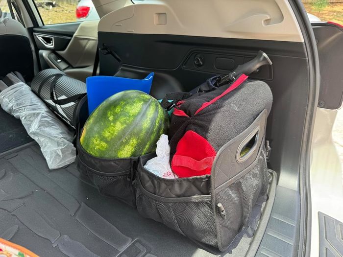 A car trunk filled with a watermelon, a red-and-black duffle bag, a blue plastic bin, and other miscellaneous items. No people are visible in the image.