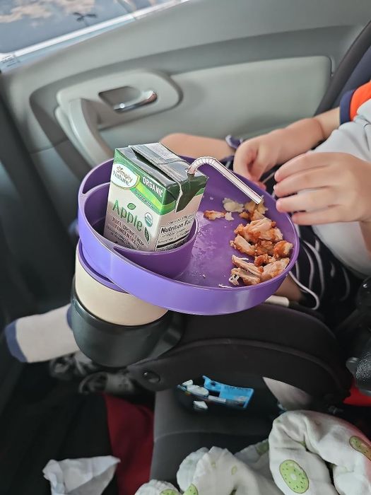 A child in a car seat uses a purple tray to hold a juice box and snacks.