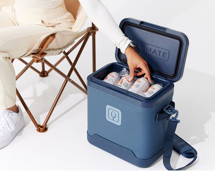 A person dressed in light clothing sitting in a chair, reaching into a BrüMate cooler filled with canned beverages.