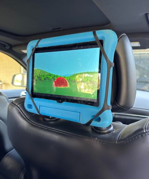 A tablet with a blue case attached to a car headrest, showcasing a vibrant animated scene with a red object and green trees.