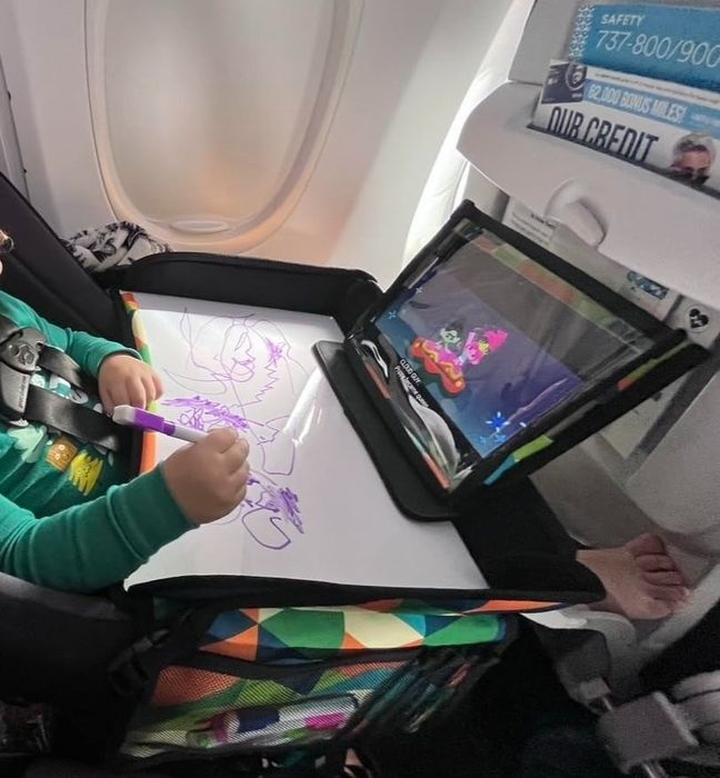 A child seated on an airplane, drawing on a whiteboard and watching a tablet placed on a tray table.