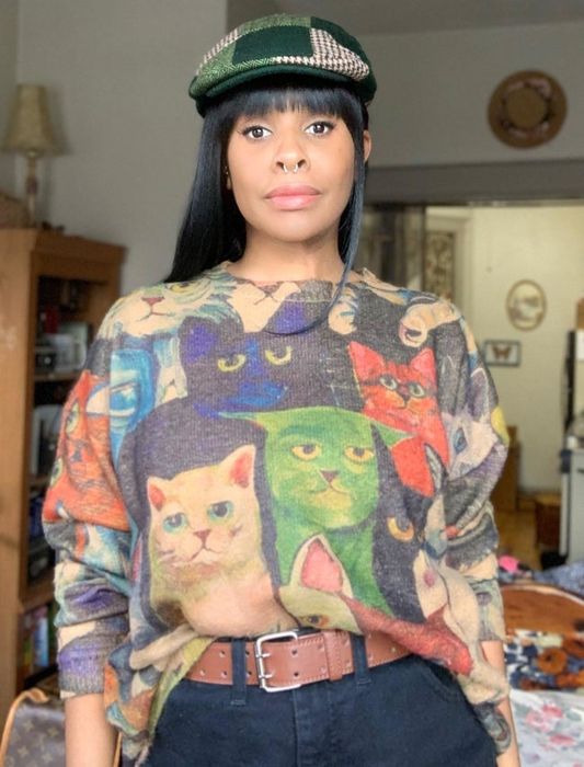 A reviewer styled in the cat-themed graphic sweater, tucked into black jeans with a brown belt