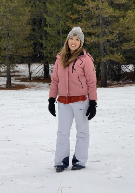 A reviewer models snow pants while standing in a snowy landscape.