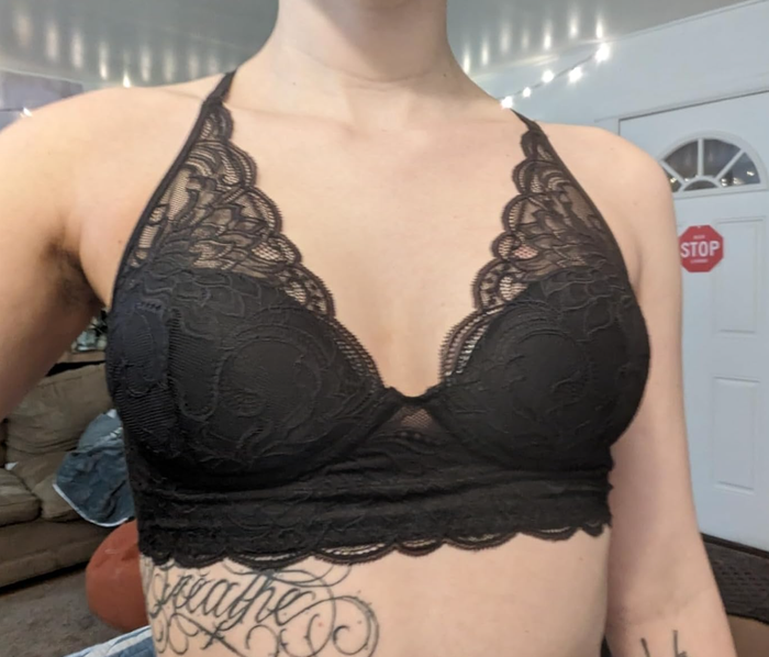 A person models a black lace bralette with detailed designs, showcasing tattoos on their chest and left arm