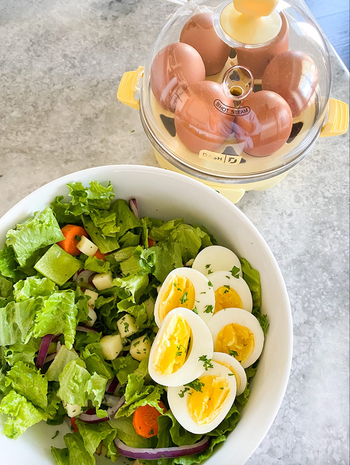 A dome-shaped yellow egg cooker filled with perfectly cooked eggs, placed beside a salad garnished with hard-boiled eggs