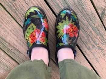 A review image featuring black clogs with green leaves and red flowers, displayed on a wooden deck.