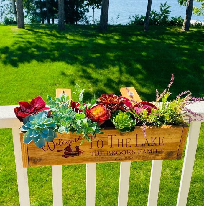 A fence adorned with plants and a personalized sign that reads 'Welcome to the Lake, The Brooks Family,' with a scenic lake in the background.