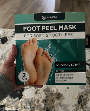 A hand holding a box of Dermora Foot Peel Mask, which includes two pairs for exfoliating feet.