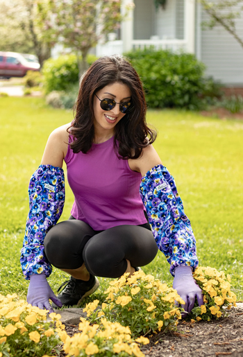 A woman wearing sunglasses and a floral-sleeved top, gardening among vibrant yellow flowers.
