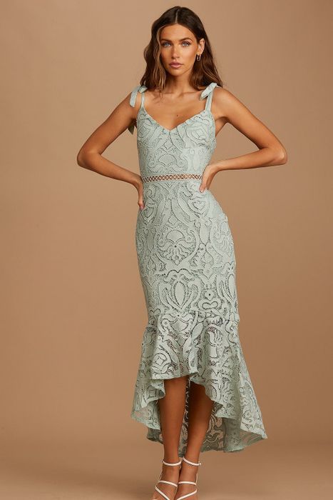 A woman posing in a light green lace dress, featuring a high-low hemline and delicate spaghetti straps