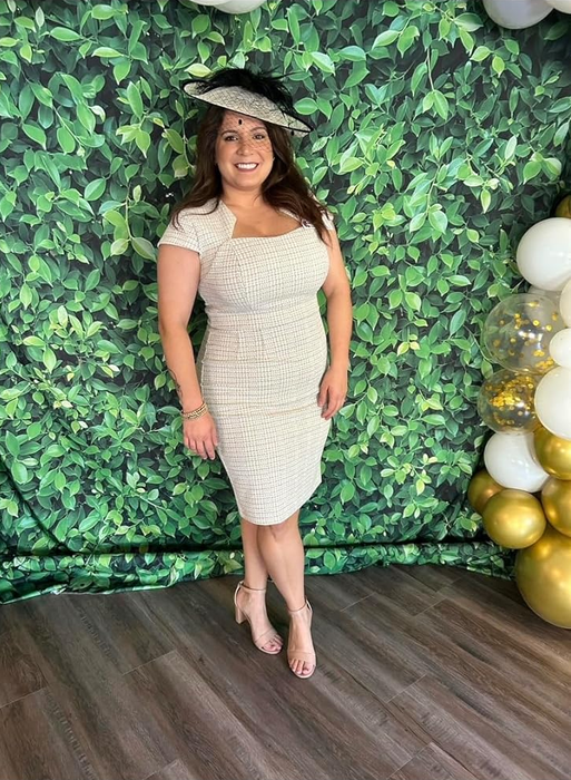 A woman in a beige dress and hat posing in front of a leafy background adorned with gold and white balloons.