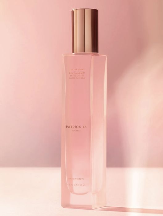 A bottle of Patrick Ta Major Glow Dewy Milk Mist with a gold cap, placed on a flat surface against a muted backdrop