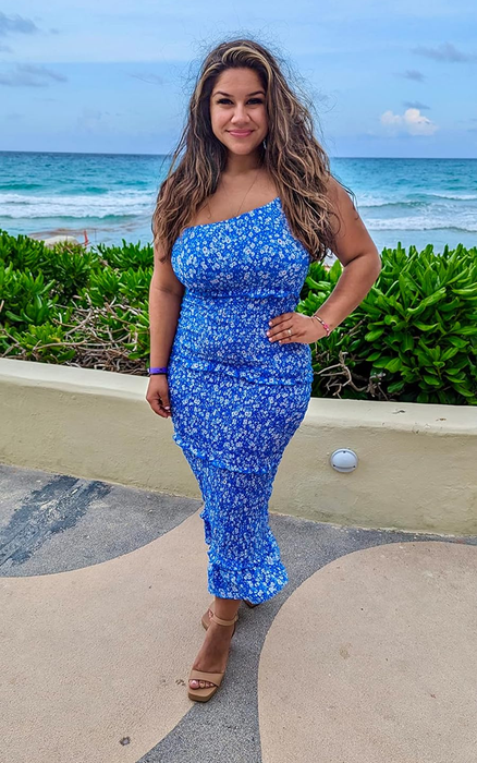 Reviewer in a blue floral dress posing by the beach with one hand on her hip