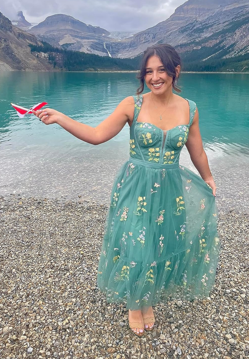 A reviewer in an embroidered dress posing in a serene setting with water and mountains, holding a paper plane.