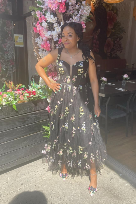 A reviewer wearing an elegant evening dress adorned with floral appliqué, standing gracefully in front of a beautifully decorated flower-covered entrance.