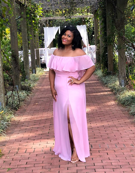 A reviewer posing outdoors in an off-shoulder pink gown with ruffled details and a leg slit.