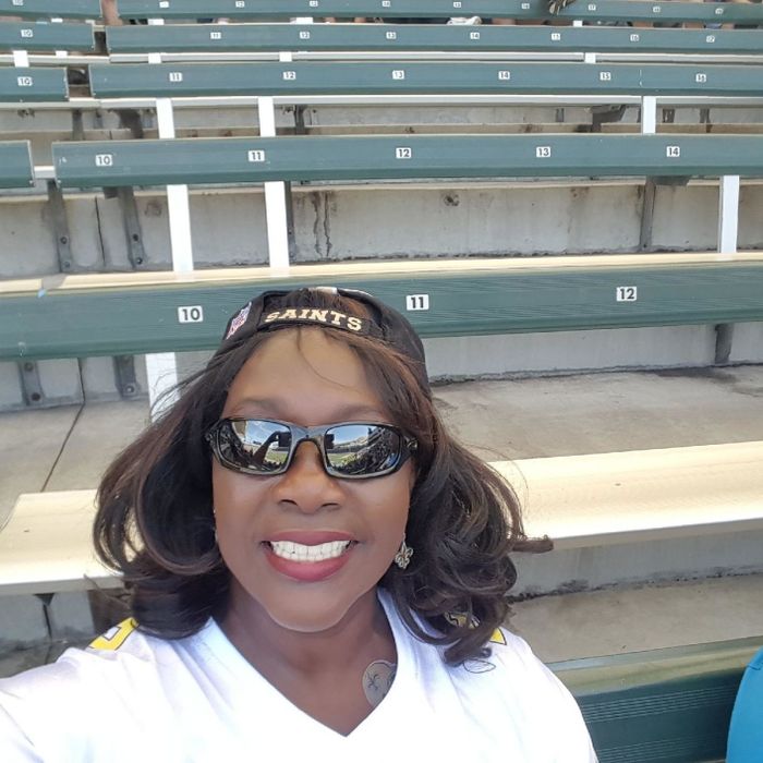 A woman wearing a 'Saints' cap and sunglasses smiles in a selfie taken at a nearly empty stadium