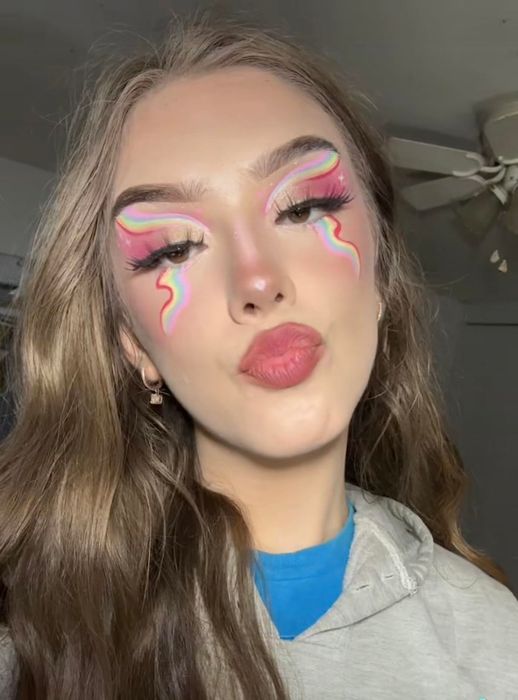 A critic with lengthy locks dressed in a hoodie, sporting dramatic eye makeup featuring vibrant pink hues and rainbow patterns radiating from their eyes.