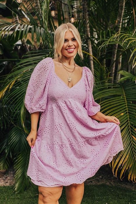 A woman is smiling and posing in a v-neck eyelet dress, hands confidently placed on her hips.