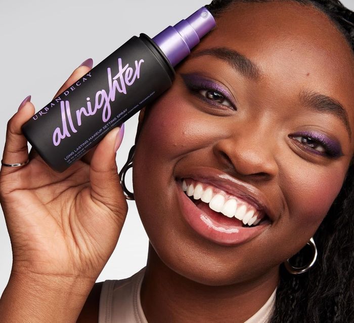 A model smiling while holding an Urban Decay All Nighter setting spray near their face
