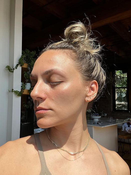 A reviewer, eyes shut and hair casually tied up in a messy bun, dons a camisole paired with a subtle necklace, captured in a room bathed in natural sunlight
