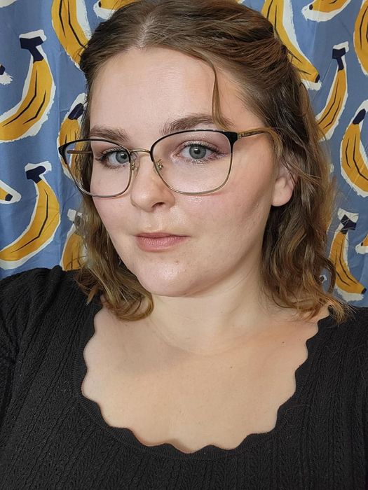 The individual in the photo, whose name is not mentioned, is wearing glasses and a black scalloped top, smiling against a backdrop with a banana print
