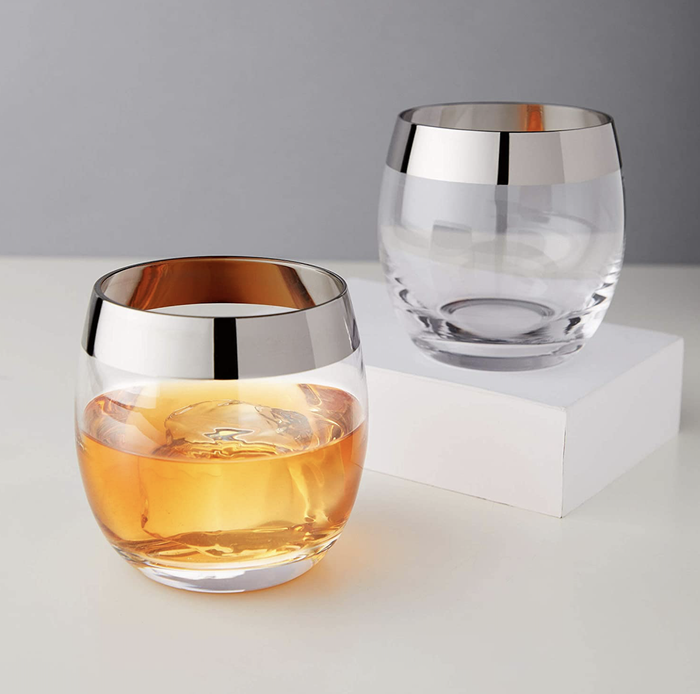 One glass, half-filled with liquor and featuring a chrome rim, alongside another empty glass, also rimmed in chrome.