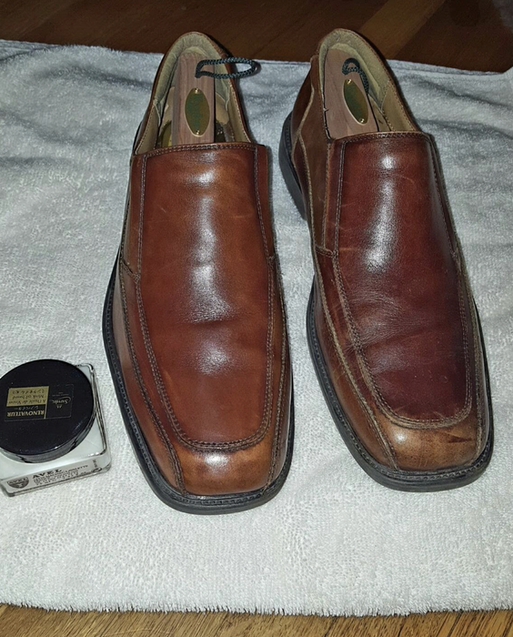 A jar of leather conditioner placed beside a pair of brown leather shoes, with the shoe on the left showing more vibrancy and fewer scuffs after being treated with the conditioner.