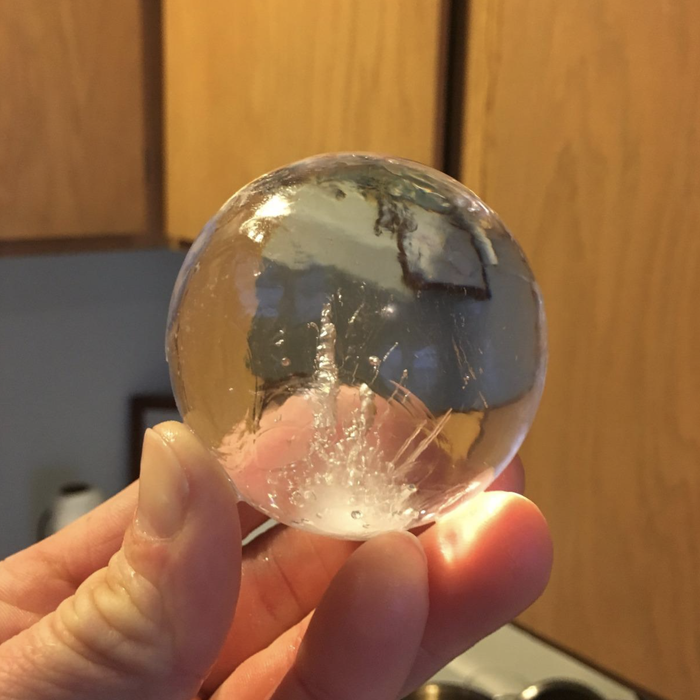 A hand gently holding a clear ice sphere, gripped with the tips of the fingers and no silicone mold in sight.